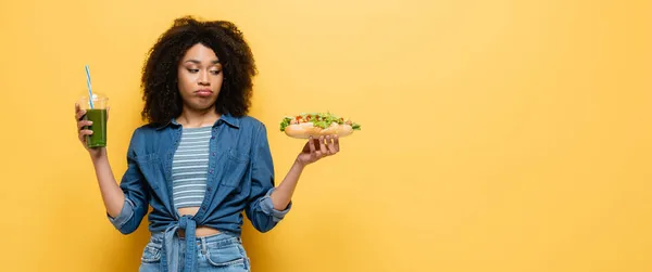 Confused african american woman looking at hot dog while holding fresh smoothie on yellow, banner — Stock Photo