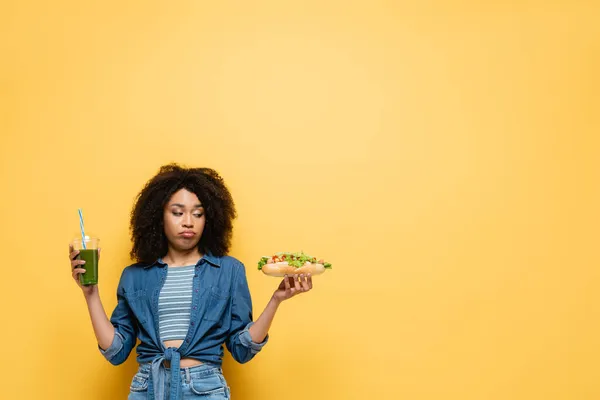 Displeased african american woman choosing between hot dog and fresh smoothie on yellow — Stock Photo