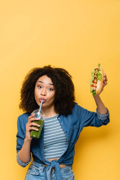 Afroamerikanerin trinkt frischen Smoothie, während sie Hot Dog auf gelb hält — Stockfoto
