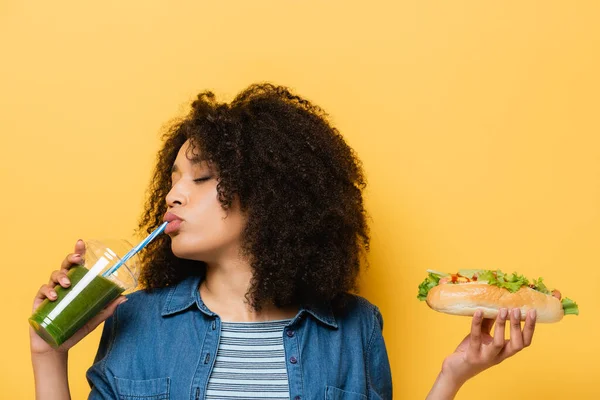 African american woman with closed eyes drinking fresh smoothie while holding hot dog isolated on yellow — Stock Photo