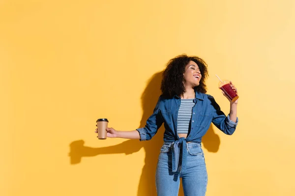 Joyful african american woman posing with fresh smoothie and coffee to go on yellow — Stock Photo