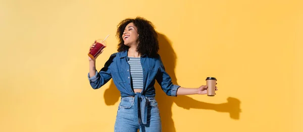 Mujer afroamericana complacida sosteniendo café y batido fresco en amarillo, pancarta - foto de stock