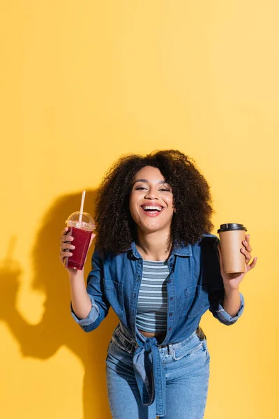 Fröhliche afrikanisch-amerikanische Frau mit Kaffee und frischem Smoothie lächelt in die Kamera auf gelb — Stockfoto