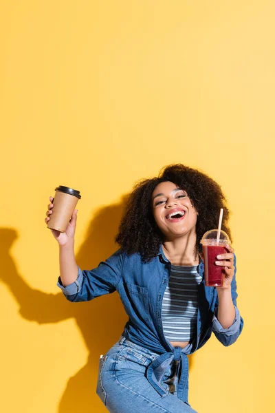 Mujer afroamericana emocionada con batido y café para ir mirando a la cámara en amarillo — Stock Photo