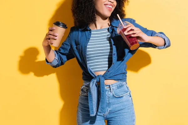 Partial view of smiling african american woman holding coffee to go and smoothie on yellow — Stock Photo