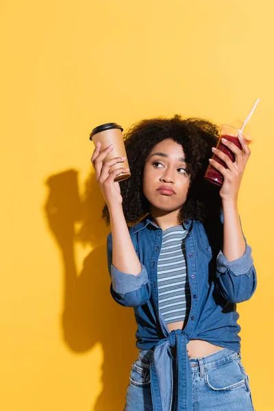 Confused african american woman choosing between coffee and fresh smoothie on yellow — Stock Photo