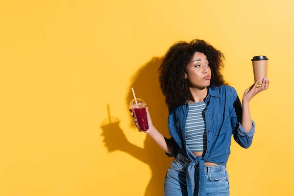 Mujer afroamericana desalentada sosteniendo batido fresco mientras mira el café para ir en amarillo — Stock Photo