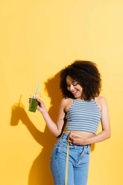 Mulher americana africana alegre medindo cintura enquanto segurando smoothie fresco no amarelo — Fotografia de Stock