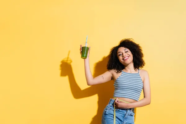 Slim and happy african american woman with fresh smoothie measuring waist on yellow — Stock Photo