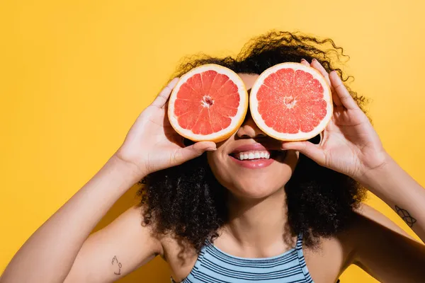 Gaie femme afro-américaine obscurcissant visage avec des moitiés de pamplemousse juteux sur jaune — Photo de stock