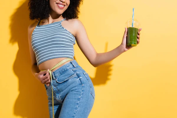 Vista parziale della donna afro-americana soddisfatta che misura la vita mentre tiene il frullato fresco sul giallo — Foto stock