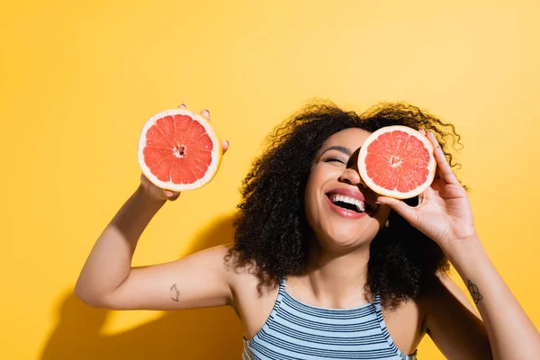 Excited african american woman laughing while holding halves of fresh grapefruit on yellow — Stock Photo