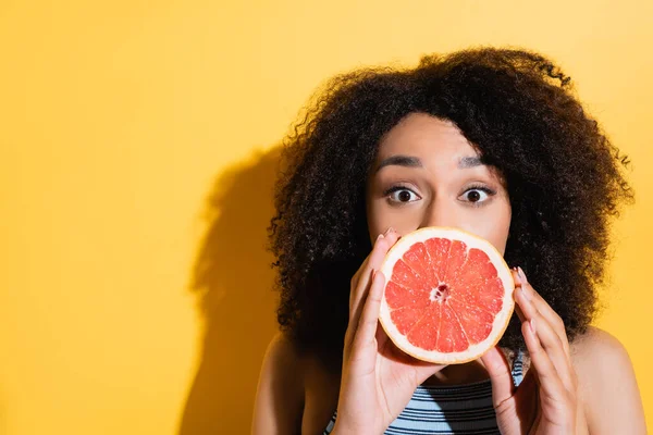 Jeune femme afro-américaine couvrant la bouche avec la moitié de pamplemousse frais sur jaune — Photo de stock