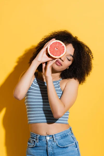 Young african american woman in striped tank top obscuring face with half of ripe grapefruit on yellow — Stock Photo