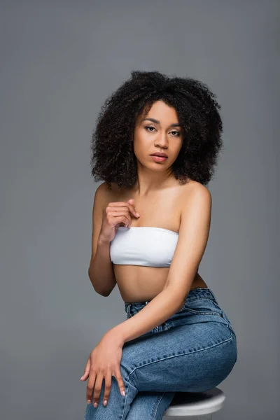 Mujer afroamericana en top blanco y jeans mirando cámara aislada en gris - foto de stock