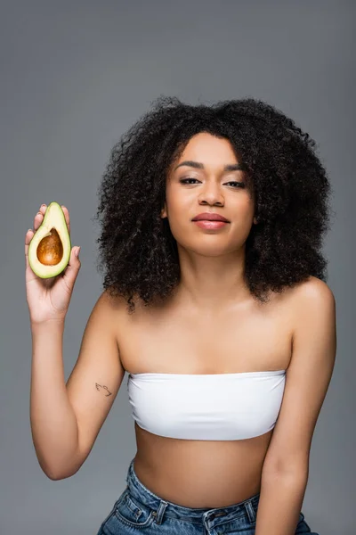 Young african american woman in white top holding half of avocado isolated on grey — Stock Photo