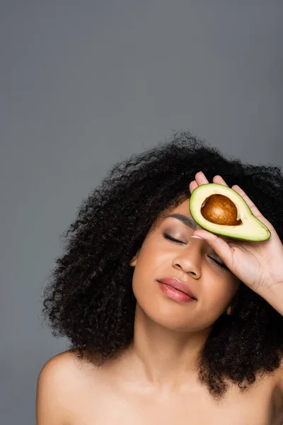 Mujer afroamericana con los ojos cerrados sosteniendo aguacate cortado cerca de la cara aislado en gris - foto de stock