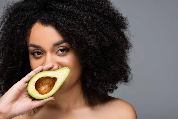 Mujer afroamericana bastante sosteniendo la mitad de aguacate fresco cerca de la cara aislado en gris - foto de stock