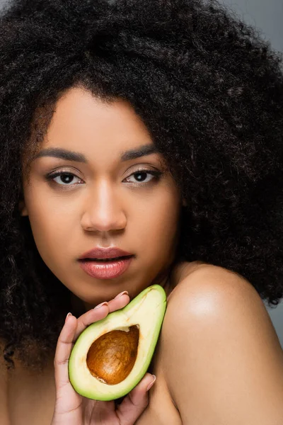 Pretty african american woman with natural makeup and perfect skin holding cut avocado isolated on grey — Stock Photo