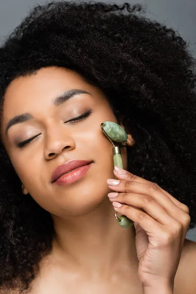 Close up view of african american woman with closed eyes using jade roller on face isolated on grey — Stock Photo