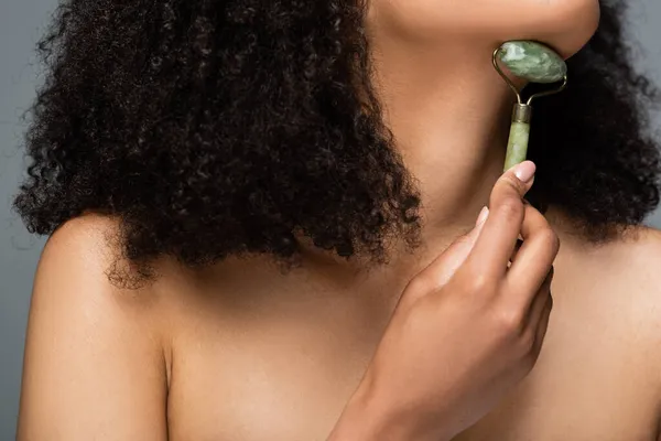 Cropped view of african american woman massaging chin with jade roller isolated on grey — Stock Photo