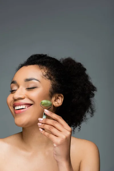 Mujer afroamericana feliz con piel perfecta y maquillaje natural sosteniendo rodillo de jade aislado en gris - foto de stock