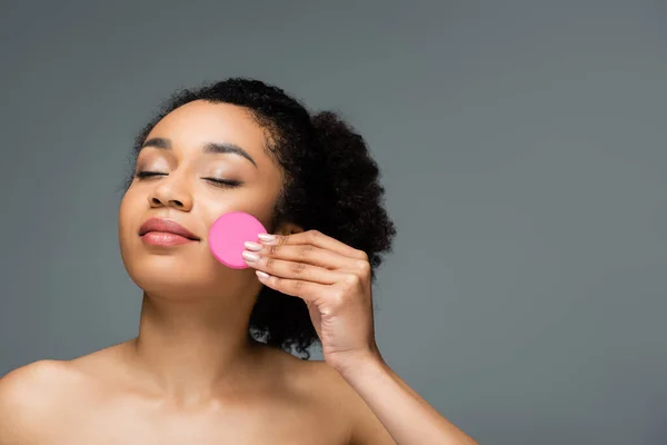 Giovane donna afroamericana con spalle nude e occhi chiusi applicando fondotinta viso con spugna isolata su grigio — Foto stock