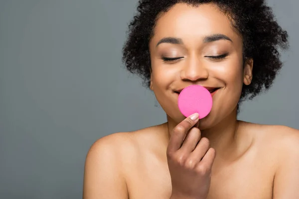 Cheerful african american woman with closed eyes and naked shoulders holding cosmetic sponge near lips isolated on grey — Stock Photo