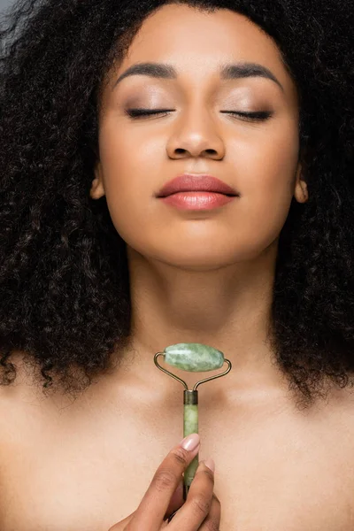 Mujer afroamericana bonita con los ojos cerrados y maquillaje natural sosteniendo rodillo de jade - foto de stock