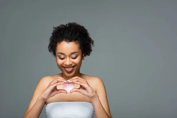 Happy african american woman holding heart-shaped cosmetic sponge isolated on grey — Stock Photo