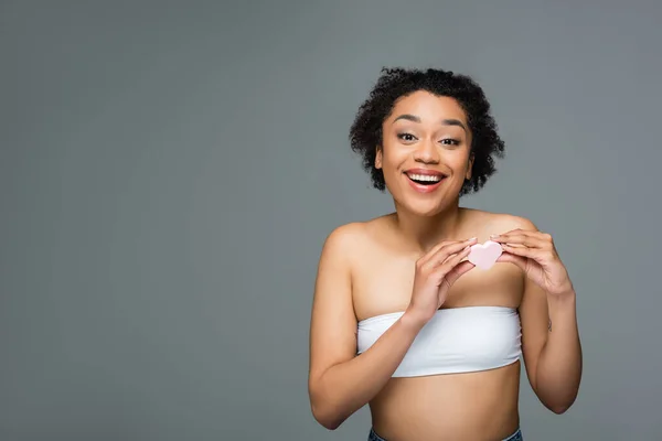 Eccitata donna afro-americana guardando la fotocamera mentre tiene la spugna cosmetica a forma di cuore isolata sul grigio — Foto stock