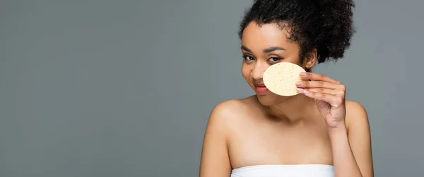 Alegre afroamericana mujer celebración cosmética esponja cerca de la cara aislado en gris, pancarta - foto de stock