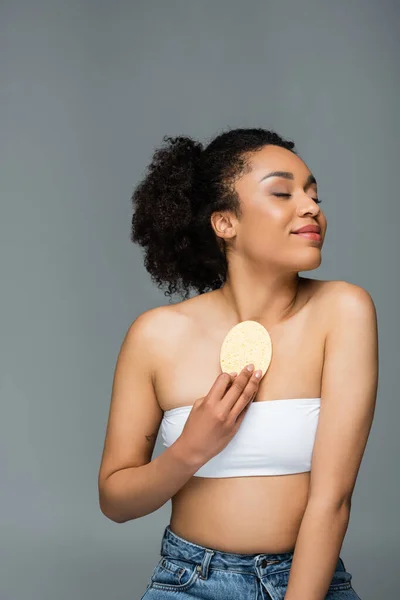 Pleased african american woman with perfect skin wiping body with cosmetic sponge isolated on grey — Stock Photo