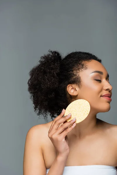 Pleased african american woman with closed eyes wiping face with cosmetic sponge isolated on grey — Stock Photo