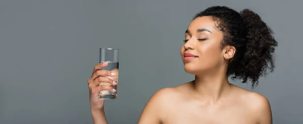 Mujer afroamericana complacida con los ojos cerrados y los hombros desnudos sosteniendo un vaso de agua dulce aislado en gris, pancarta - foto de stock