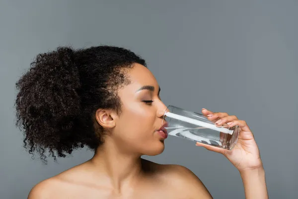 Pretty african american woman with clean skin and naked shoulders drinking water isolated on grey — Stock Photo