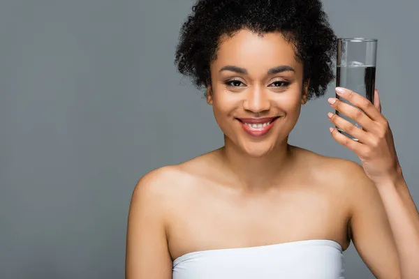 Happy african american woman with natural makeup on perfect face holding glass of water isolated on grey — Stock Photo