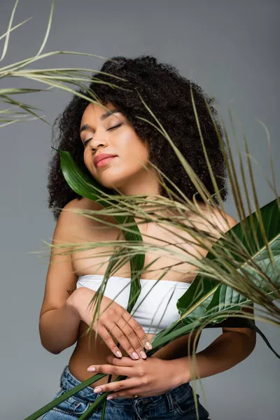 Jovem afro-americana posando com olhos fechados perto de folhas verdes isoladas em cinza — Fotografia de Stock