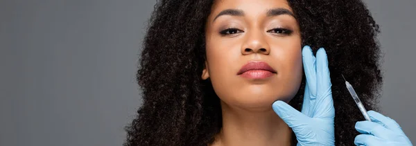 Esteticista em luvas de látex segurando injeção de beleza perto de mulher afro-americana isolada em cinza, banner — Fotografia de Stock