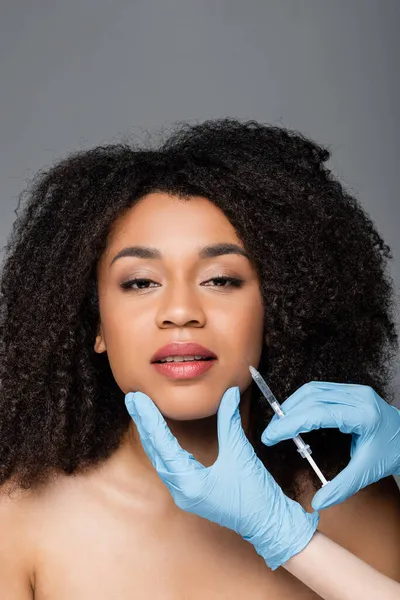 Esteticista en guantes de látex dando inyección de belleza a mujer afroamericana aislada en gris - foto de stock