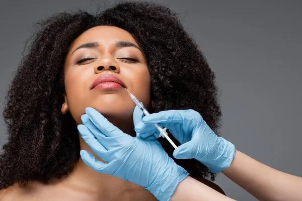 Cosmetólogo en guantes de látex haciendo inyección de belleza a labios de mujer afroamericana aislada en gris - foto de stock