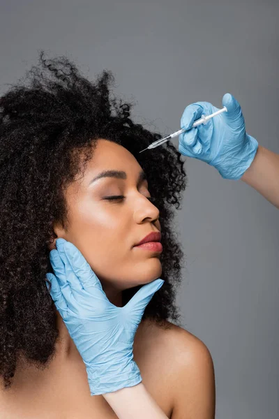 Kosmetikerin in Latex-Handschuhen macht Schönheitsinjektion für Afroamerikanerin isoliert auf grau — Stockfoto