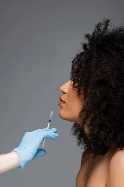 Vista lateral de mujer afroamericana cerca de cosmetólogo con inyección de belleza aislada en gris - foto de stock