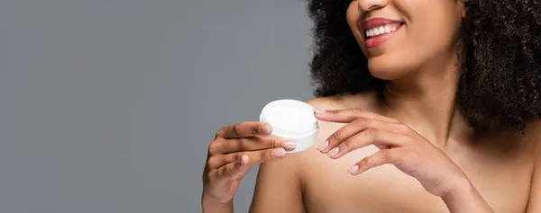 Partial view of smiling african american woman with naked shoulders holding cosmetic cream isolated on grey, banner — Stock Photo