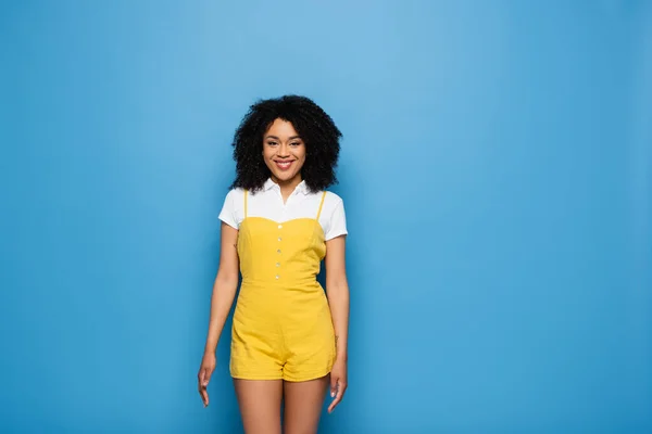 Femme afro-américaine à la mode en combinaison jaune souriant à la caméra sur bleu — Photo de stock