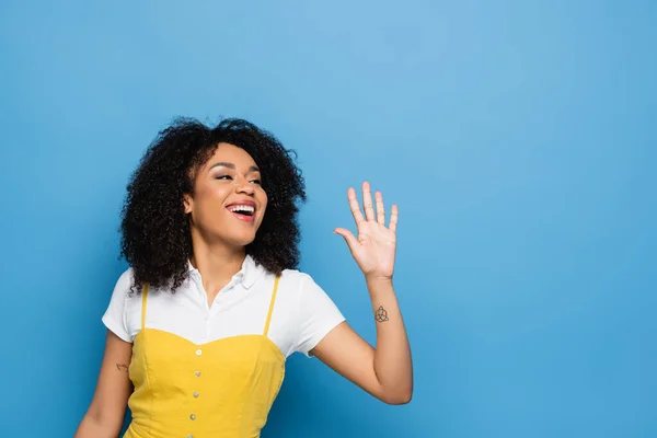 Возбужденная и счастливая африканская американка смотрит в сторону и машет рукой на синий — стоковое фото