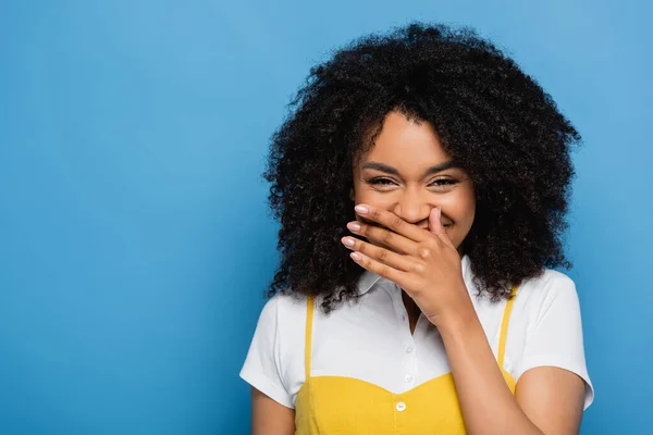 Eccitata donna afroamericana che copre la bocca con la mano mentre ride isolata sul blu — Foto stock