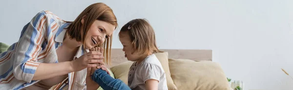 Smiling mother touching leg of child with down syndrome on bed, banner — Stock Photo