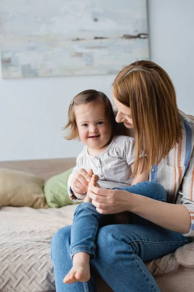 Mother touching leg of smiling daughter with down syndrome on bed — Stock Photo