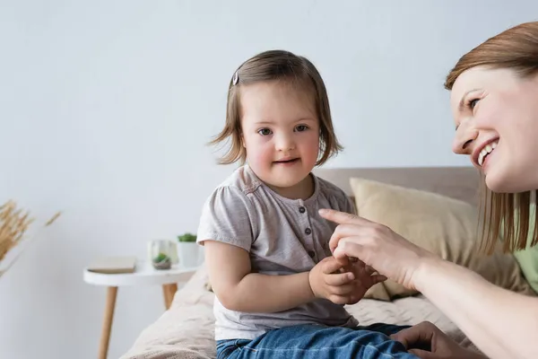 Sonriente madre señalando con el dedo a su hija pequeña con síndrome de Down en el dormitorio - foto de stock